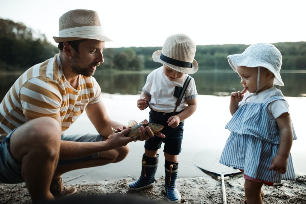 A mature father with small toddler children fishing by a lake, holding a fish.